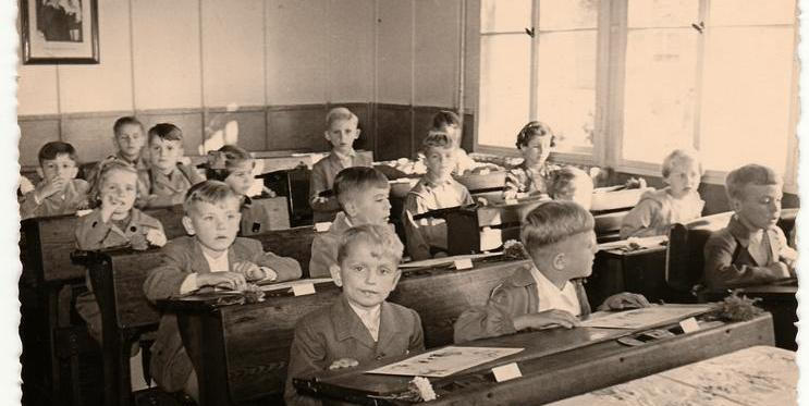 ČESKOSLOVENSKÁ SOCIALISTICKÁ REPUBLIKA – cca 50. léta: Retro fotografie zachycuje žáky sedící v dřevěných lavicích ve třídě. Dobový snímek skupiny spolužáků ve školní třídě.