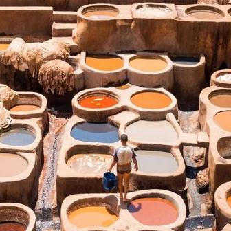 Maročan s kbelíkem mezi tradičními kamennými káděmi s přírodními barvivy, kde se zpracovávají zvířecí kůže. V minulosti zpracování kůže a textilií zahrnovalo používání hnoje, moči a jiných organických látek k měkčení a barvení materiálů. Pracovníci trávili hodiny v toxických a páchnoucích lázních bez ochranných prostředků, což způsobovalo chronické zdravotní problémy.