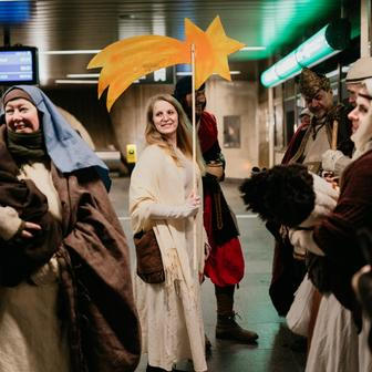 Na eskalátoru se pomalu pohybují postavy v kostýmech, které jako by patřily k samotnému mechanismu stanice.