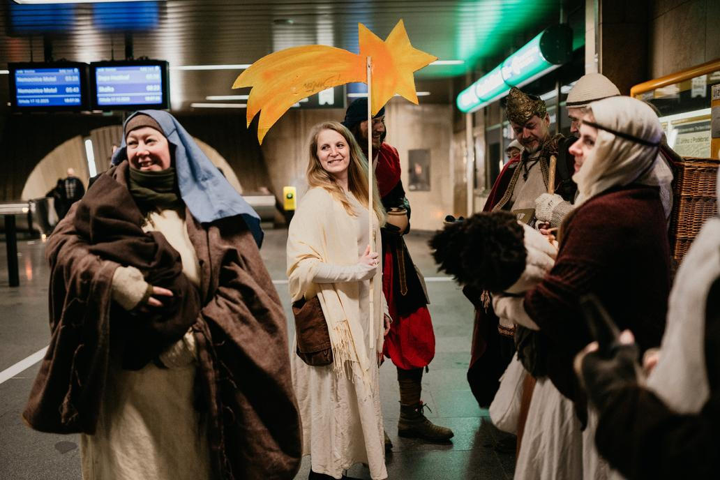 Na eskalátoru se pomalu pohybují postavy v kostýmech, které jako by patřily k samotnému mechanismu stanice.