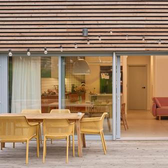 Kuchyně je řešena v otevřeném konceptu. Spodní skříňky v cihlovém tónu navazují na horní modrošedé plochy, zatímco otevřené policové moduly vizuálně odlehčují horní část sestavy a umožňují vystavit keramiku a sklo. Mobilní ostrůvek v kombinaci ocelového rámu a březové překližky na kolečkách podtrhuje flexibilitu prostoru a spolu s minimalistickými bílými barovými židlemi vnáší do interiéru lehce industriální akcent.