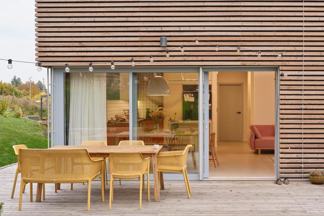 Kuchyně je řešena v otevřeném konceptu. Spodní skříňky v cihlovém tónu navazují na horní modrošedé plochy, zatímco otevřené policové moduly vizuálně odlehčují horní část sestavy a umožňují vystavit keramiku a sklo. Mobilní ostrůvek v kombinaci ocelového rámu a březové překližky na kolečkách podtrhuje flexibilitu prostoru a spolu s minimalistickými bílými barovými židlemi vnáší do interiéru lehce industriální akcent.