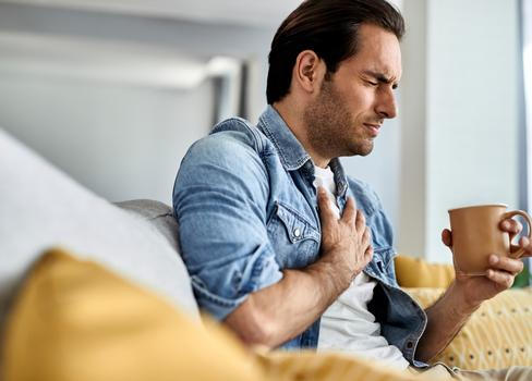 Young man feeling sick and holding his chest in pain while drinking tea in the living room.