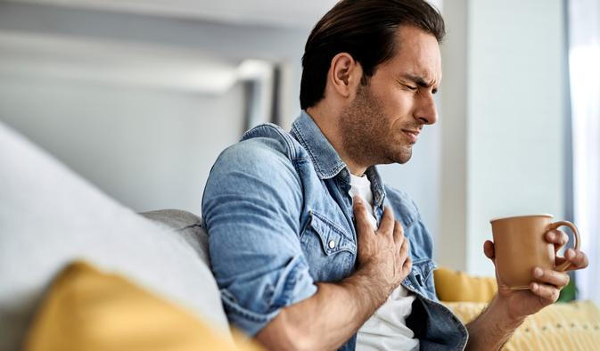 Young man feeling sick and holding his chest in pain while drinking tea in the living room.