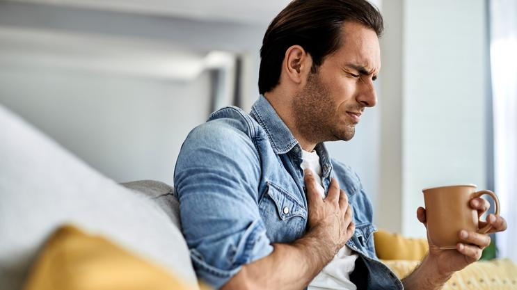 Young man feeling sick and holding his chest in pain while drinking tea in the living room.