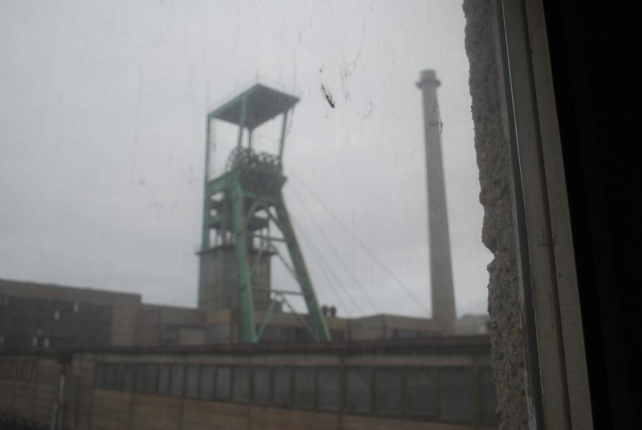 Výhled skrz zaprášené okno na těžní věž, na jejíchž lanech visí klece, které svážejí horníky pod zem.