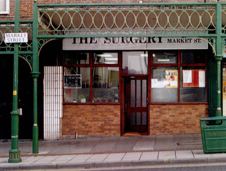 Ordinace na Market Street v Hyde, kterou si Harold Shipman otevřel v roce 1993. Místo, kde budoval pověst svědomitého rodinného lékaře — a kde nikdo netušil, že za sebou má drogovou aféru z roku 1975 ani že jeho praxe brzy přeroste hranice běžné důvěry.