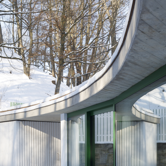 Střecha se pak rozpíná napříč celou šíří pozemku a působí jako rozepjaté křídlo od plotu k plotu. Přestože je velkorysá, nepřebíjí historickou stavbu. Naopak - původní dům zůstává hlavním aktérem. Pod severní částí střechy dále vznikl dřevěný box na zahradní náčiní, jižní část u vjezdu poskytuje kryté parkovací stání. A samotná obytná část přístavby je zasazena uprostřed za domem, přičemž díky mírnému zkosení stěn je z ulice téměř neviditelná.