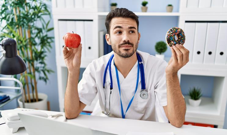 Young hispanic dietitian man holding doughnut and apple smiling looking to the side and staring away thinking. 