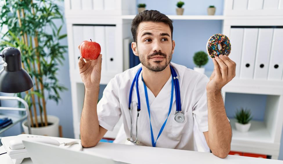 Young hispanic dietitian man holding doughnut and apple smiling looking to the side and staring away thinking. 