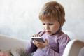 ["Indoor shot of serious sweet female child with light short hair sitting on armchair using wifi on mobile phone, having focused concentrated look while playing logical game online. Kids and gadgets"]