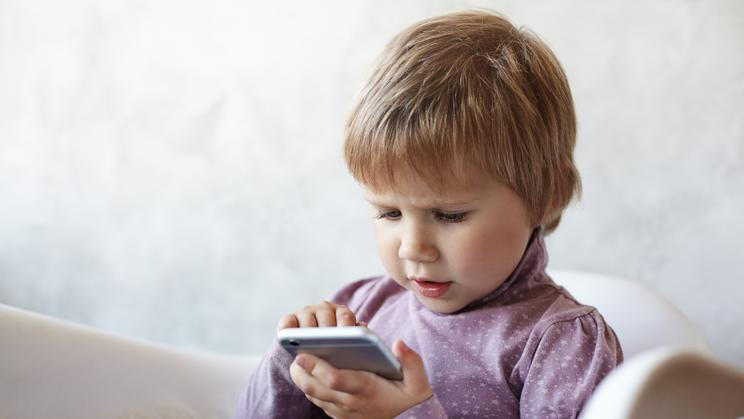 ["Indoor shot of serious sweet female child with light short hair sitting on armchair using wifi on mobile phone, having focused concentrated look while playing logical game online. Kids and gadgets"]