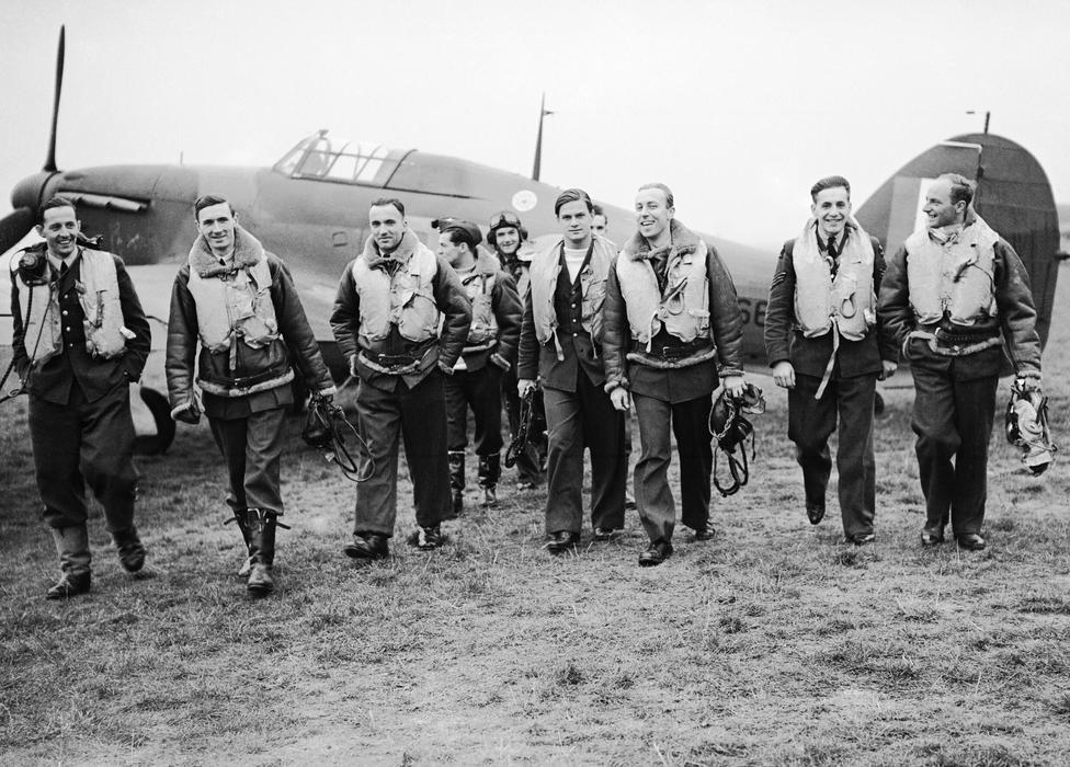 Skupina pilotů slavné polské 303. stíhací perutě kráčí od letounu Hawker Hurricane, ze kterého se podle všeho právě vrátili z bojového letu. Zleva doprava v první řadě: poručík Mirosław Ferić, kapitán John A. Kent (velitel letky „A“), nadporučík Bogdan Grzeszczak, poručík Jerzy Radomski, poručík Witold Łokuciewski, poručík Bogusław Mierzwa (zakrytý Łokuciewským), nadporučík Zdzisław Henneberg, rotmistr Jan Rogowski a rotmistr Eugeniusz Szaposznikow. Uprostřed v pozadí, v letecké kukle a brýlích, stojí poručík Jan Zumbach.