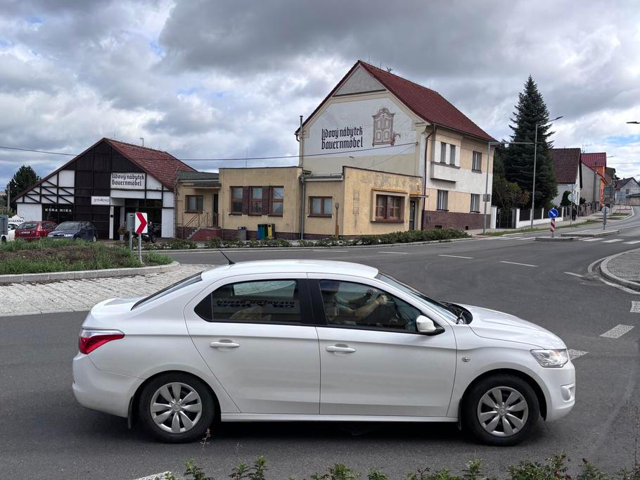 Automobil projíždí po kruhovém objezdu, za vozidlem je vidět nízká žlutá budova, v níž dříve sídlila pošta.
