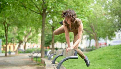 Woman performs agile parkour move over a bench in a lush green park setting.
