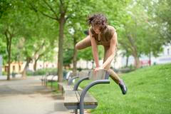 Woman performs agile parkour move over a bench in a lush green park setting.