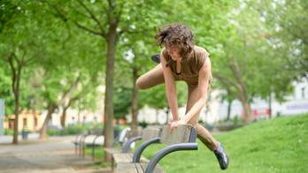 Woman performs agile parkour move over a bench in a lush green park setting.