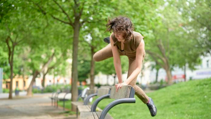 Woman performs agile parkour move over a bench in a lush green park setting.
